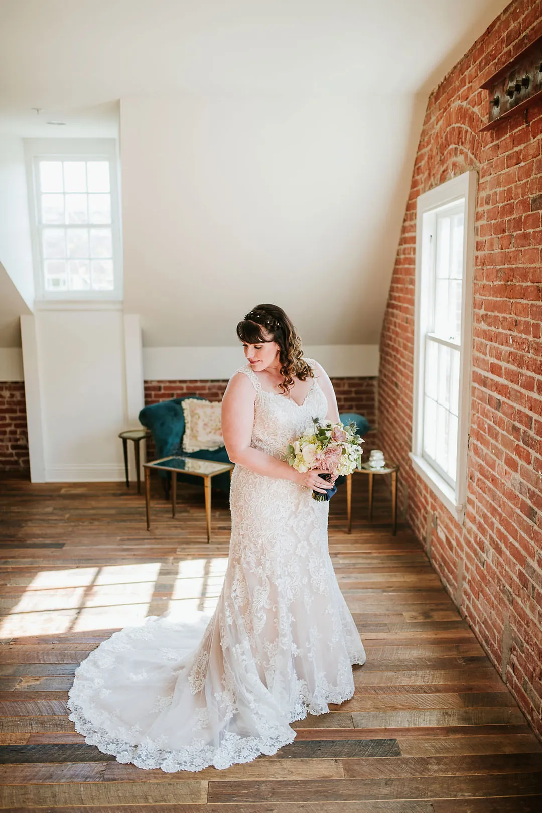 Bride stands in a sunlit loft holding a bouquet.