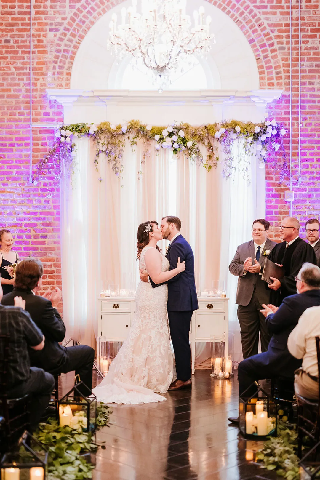 Newlyweds kiss beneath a purple lit floral backdrop.