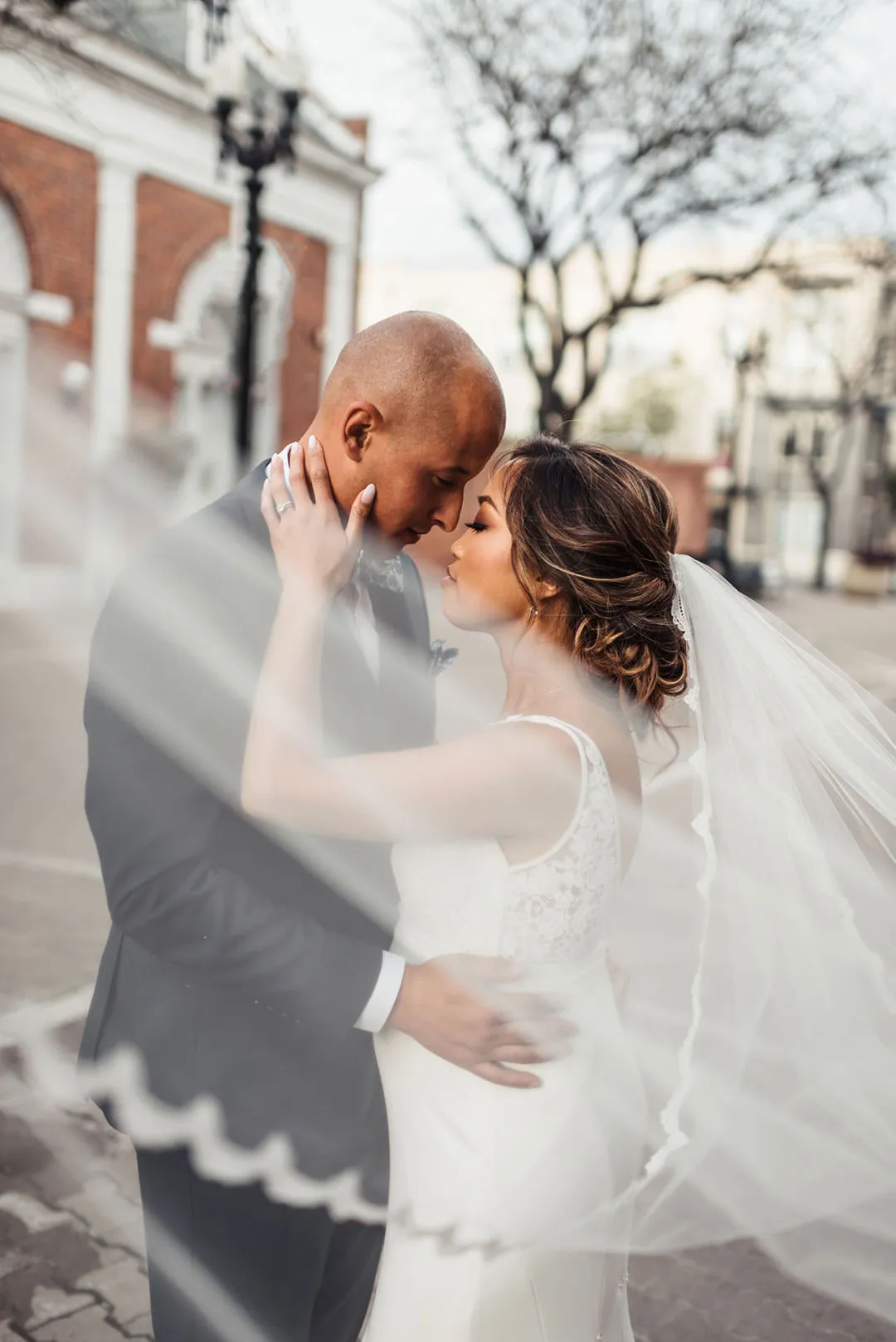 Bride and groom embrace beneath the bride’s veil on a city sidewalk.