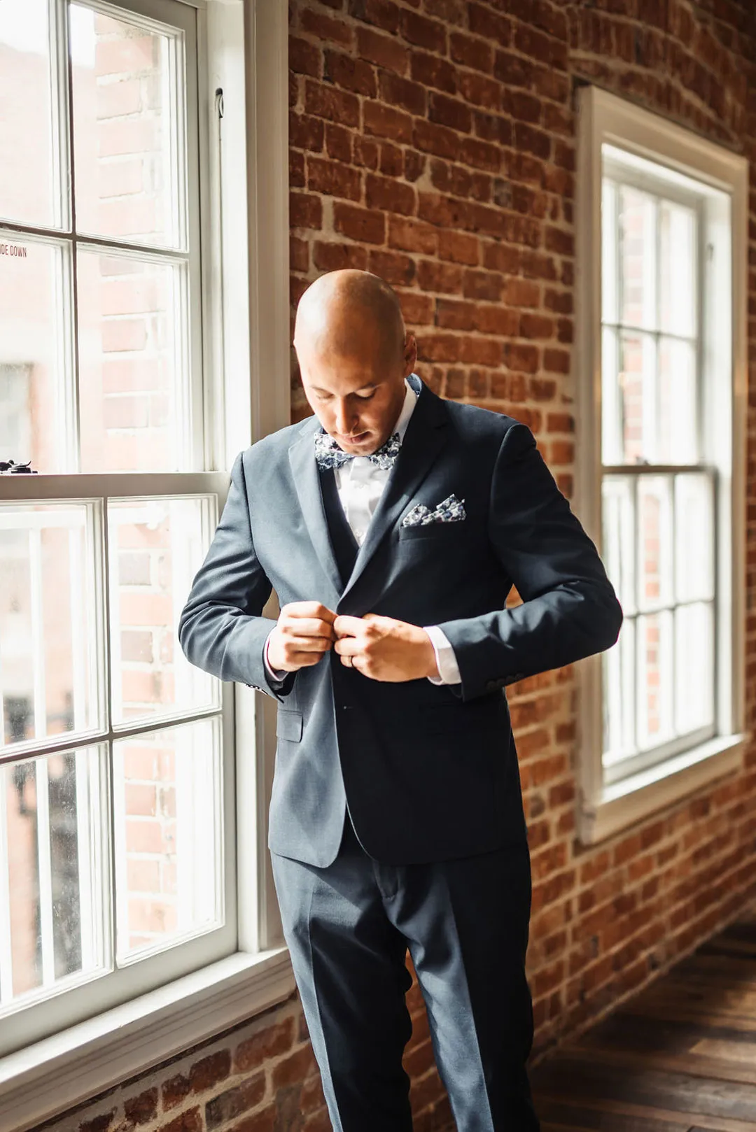 Groom buttons his dark suit beside tall windows and exposed brick.