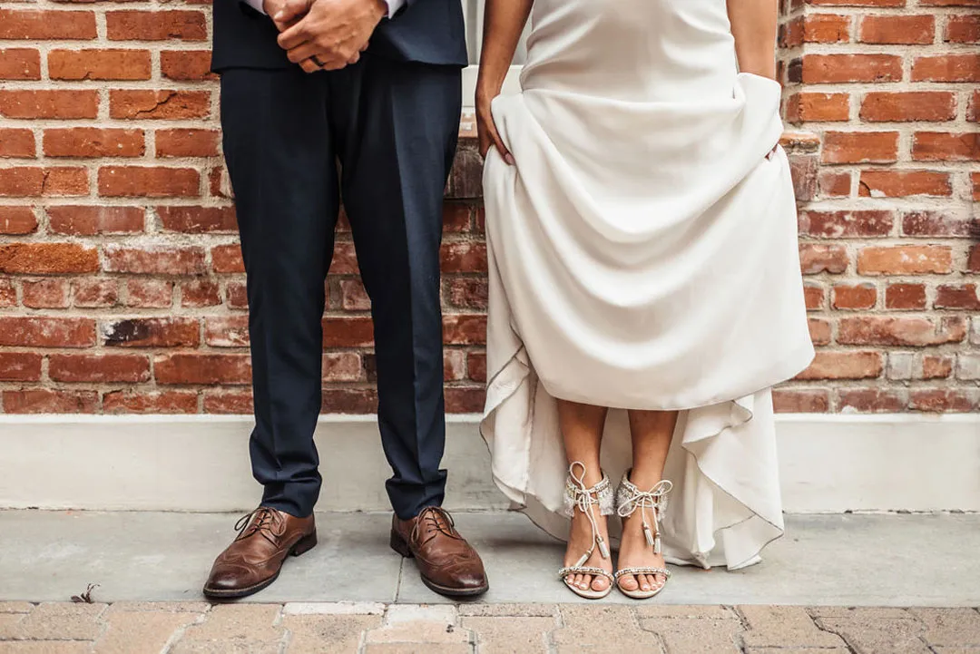 The bride’s silver heels and groom’s brown shoes beside a brick wall.
