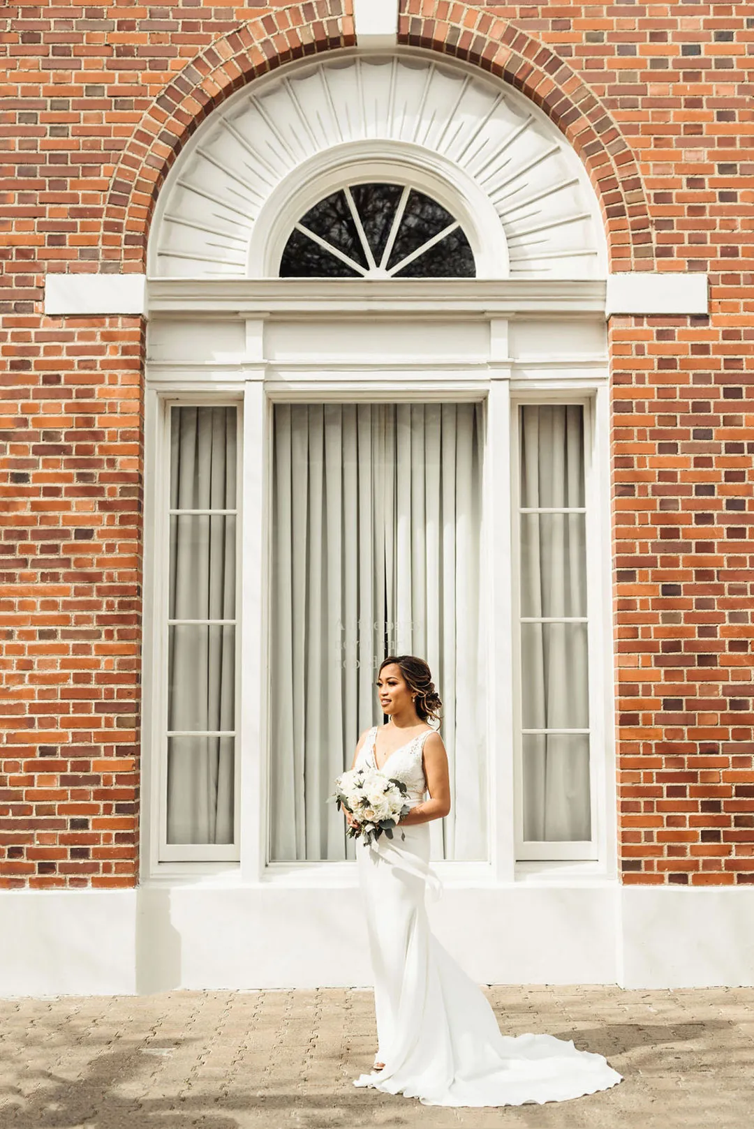Bride holds a white bouquet beneath an arched window on the brick facade.