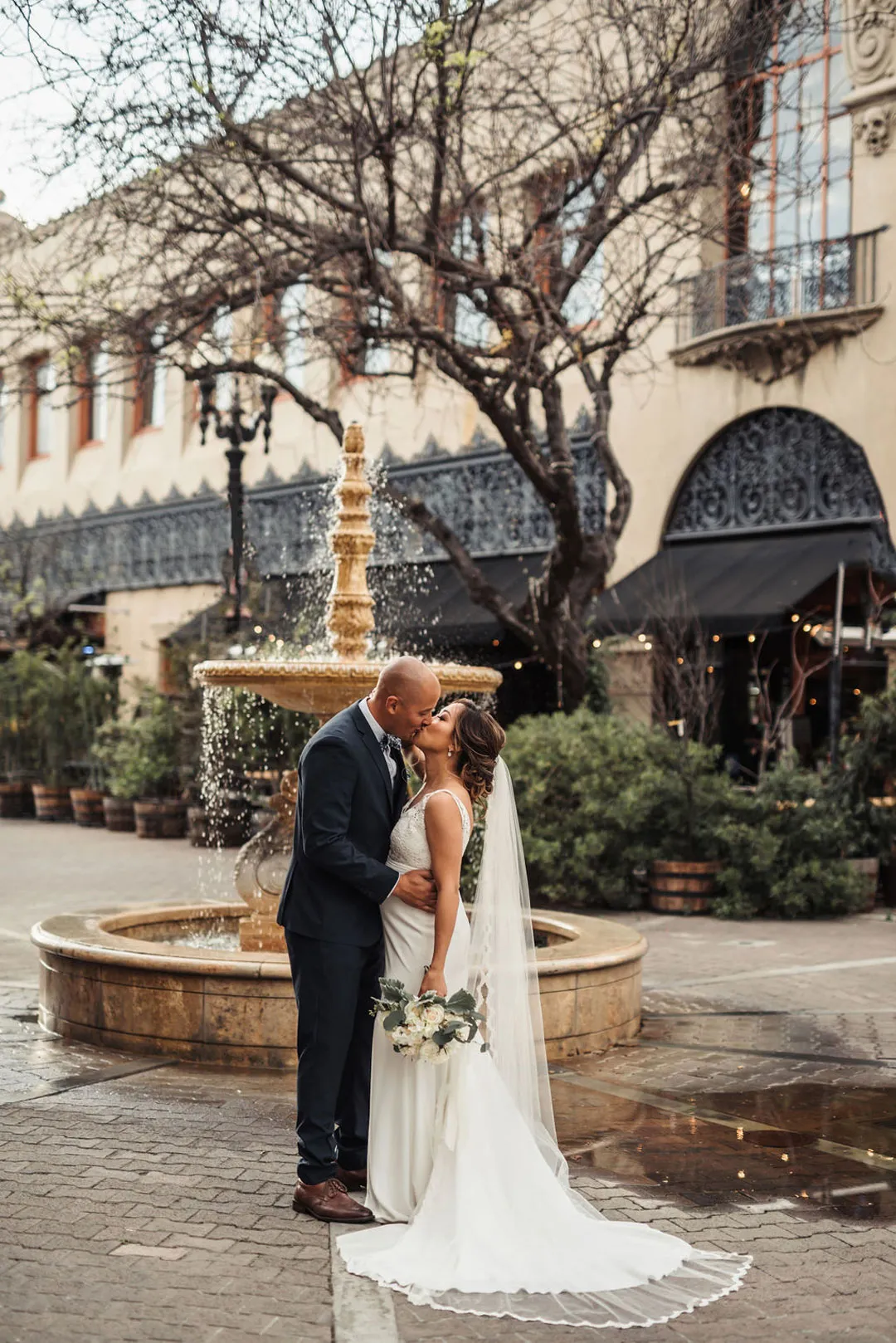 Bride and groom kiss beside a fountain in an outdoor courtyard.