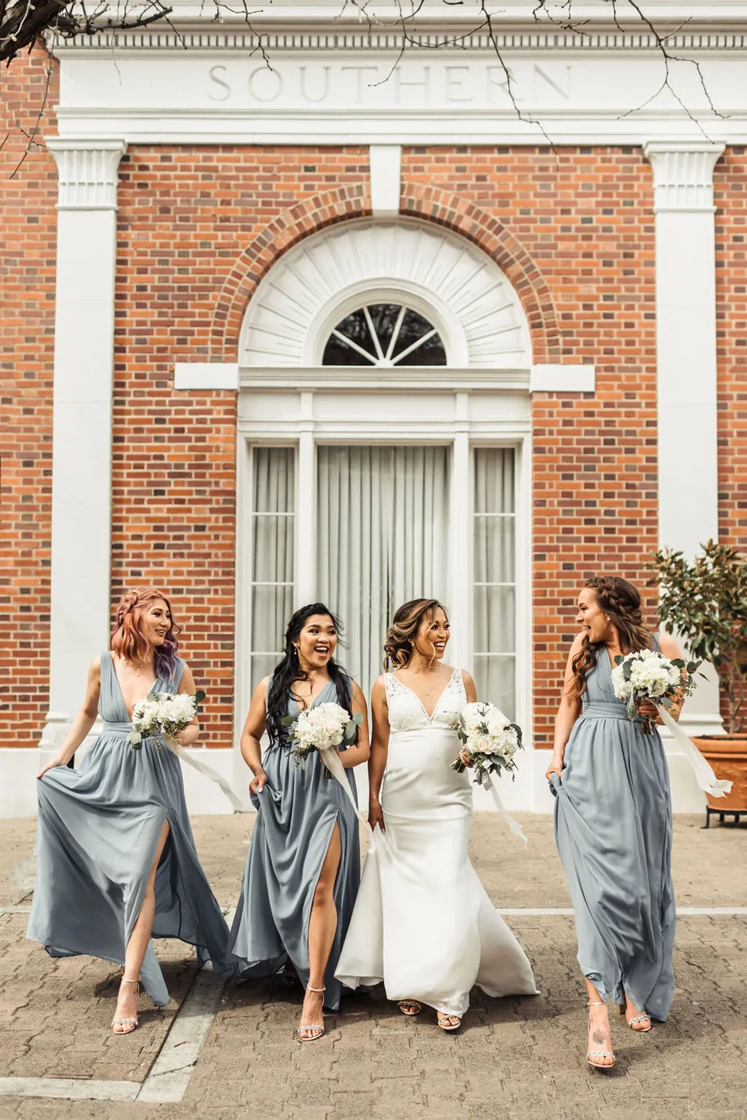 Bride walks outside with bridesmaids wearing blue-gray gowns and holding white bouquets.