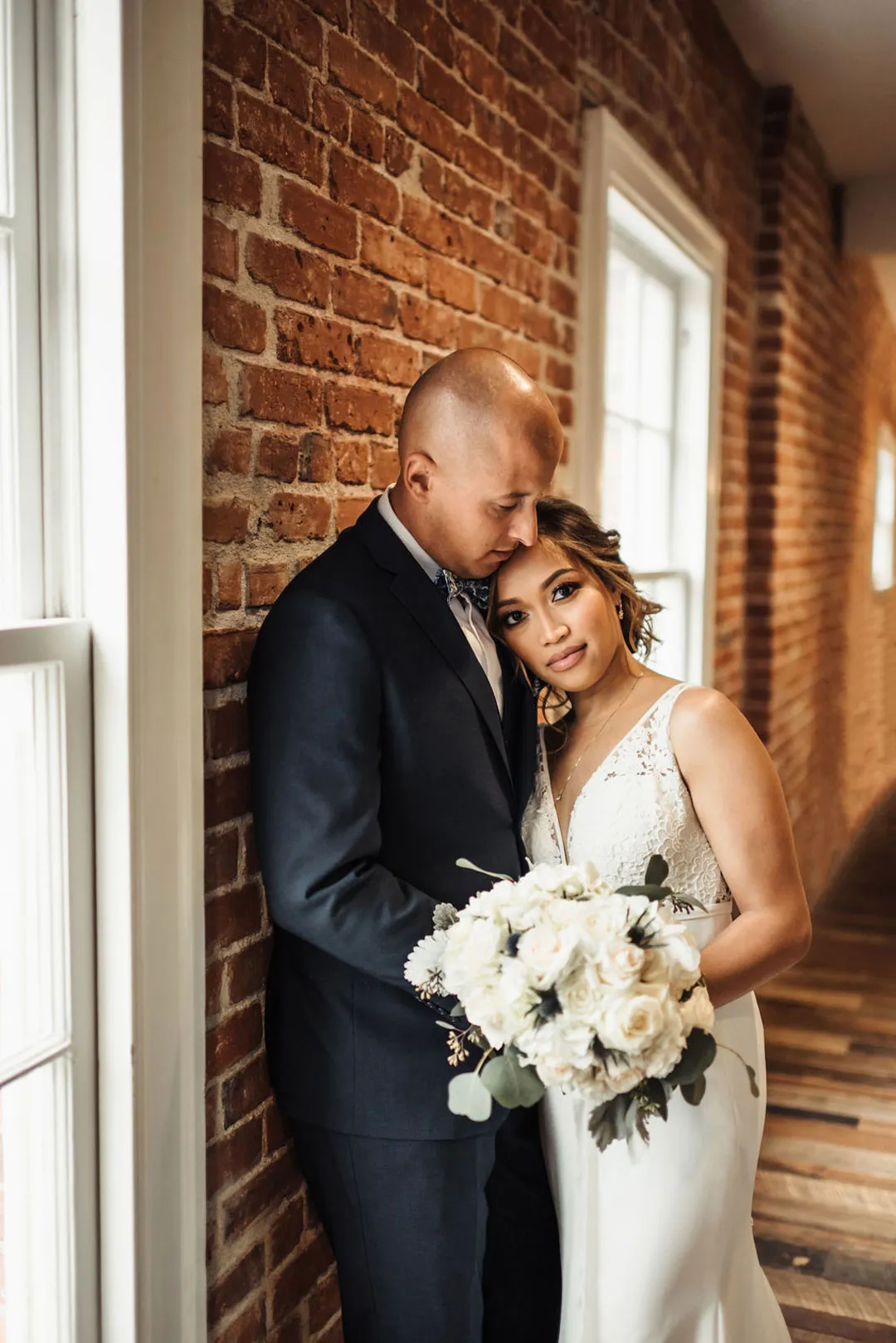 Bride and groom stand together beside a window, holding a white bouquet.