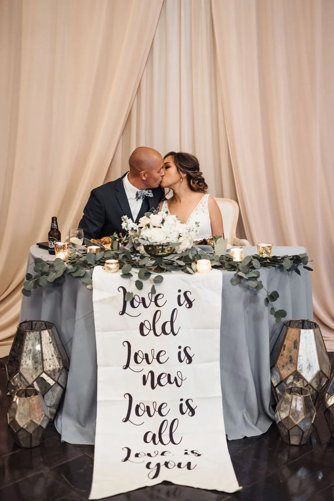 Newlyweds kiss at a sweetheart table decorated with eucalyptus and a love banner.