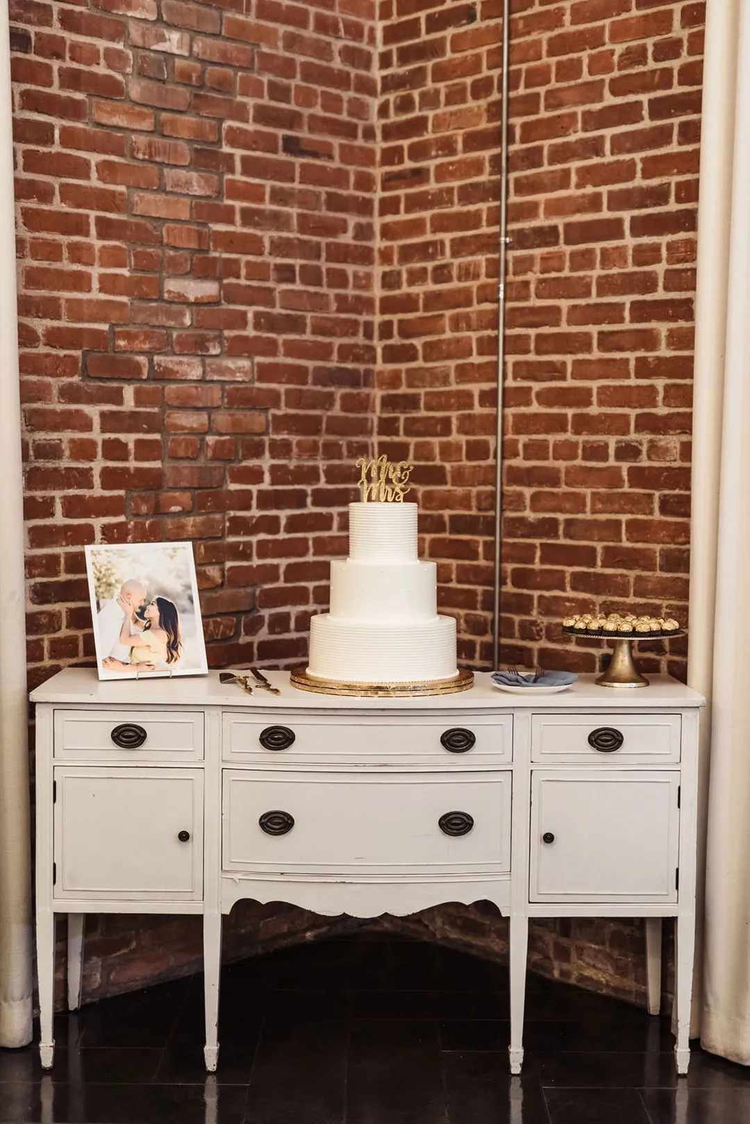 Three-tier white wedding cake on a sideboard with a framed portrait and desserts.