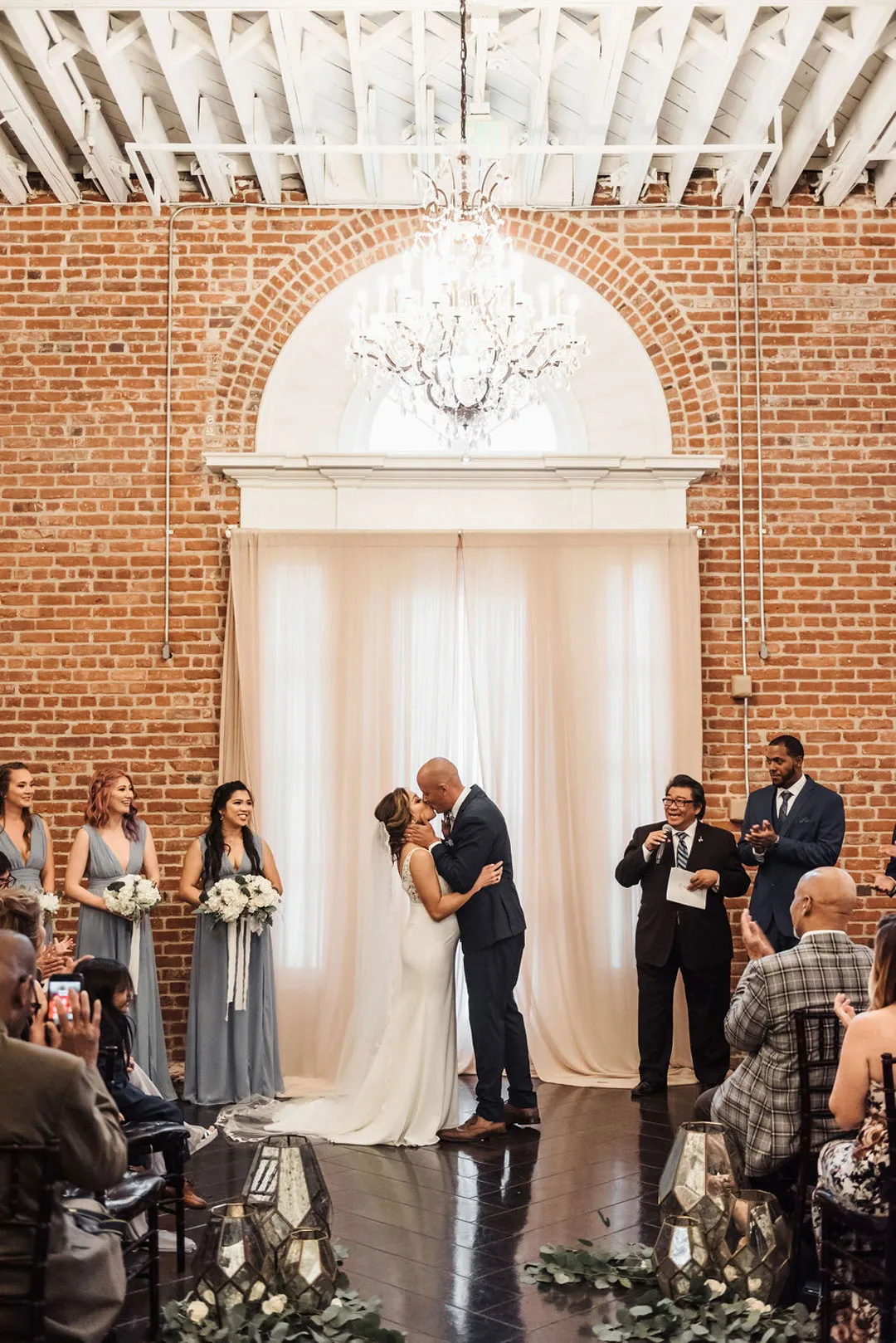 Bride and groom kiss during their ceremony beneath an arched ballroom window.