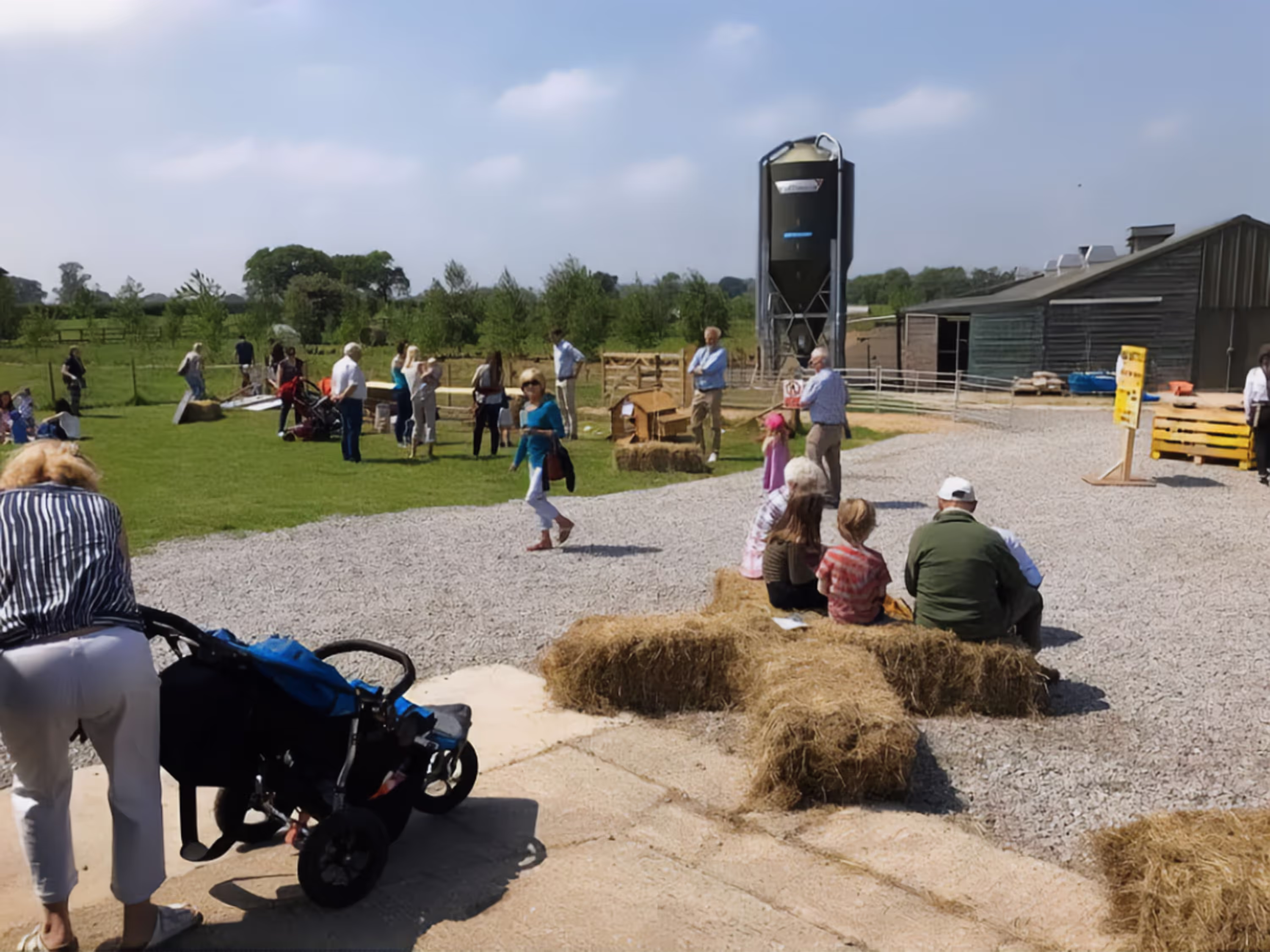People at a farm event with hay bale seating, gravel path, green field, and farm buildings.