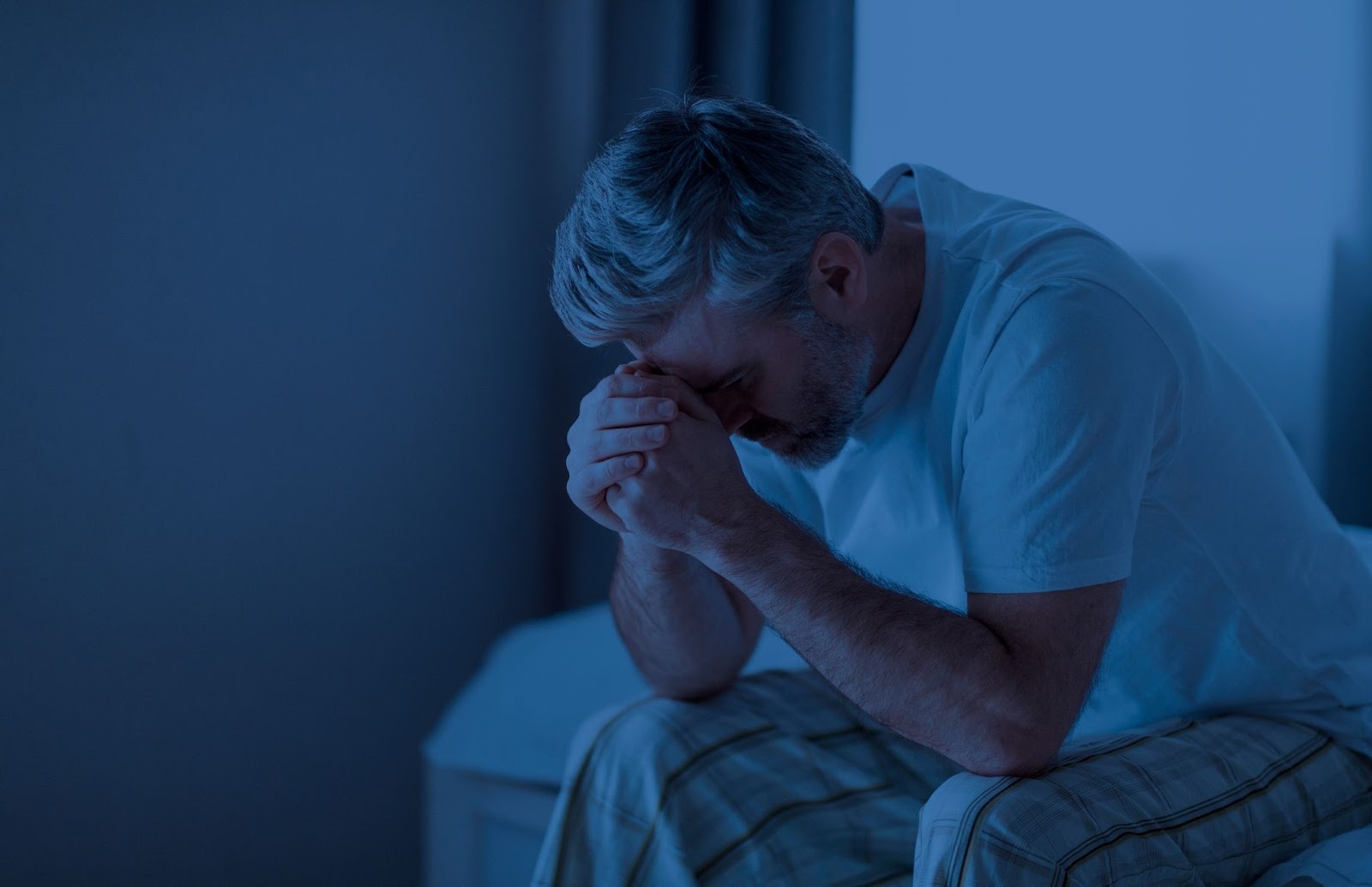 Middle-aged man awake at night with hands clasped in frustration, representing nighttime urination and sleep disruption from BPH symptoms.