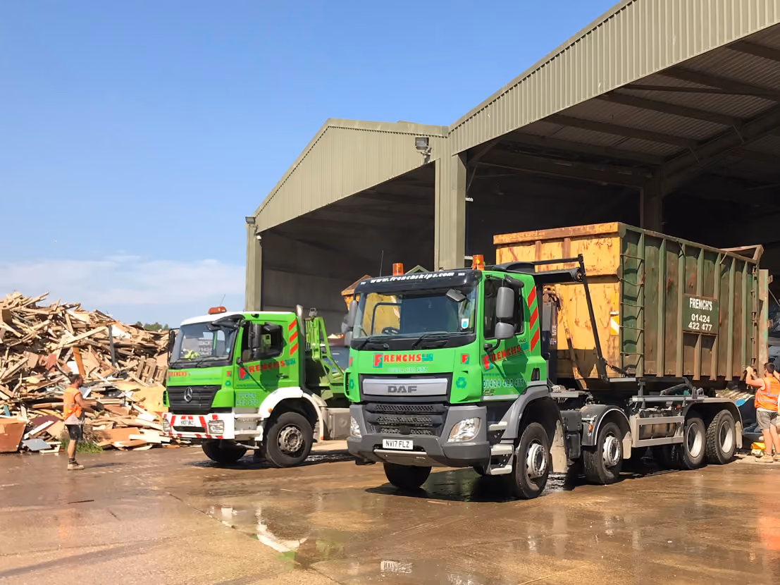 Two large green trucks parked outside a warehouse, one carrying a loaded skip.