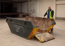 A person opens the cover of an empty metal skip bin in a concrete area.
