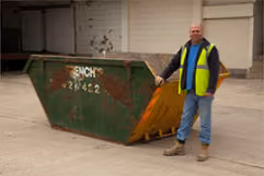 A person in a yellow safety vest stands next to a large green dumpster outside.
