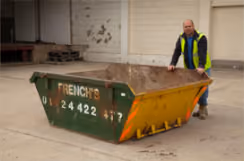 A man in a reflective vest is pushing a green and yellow skip on a concrete surface.