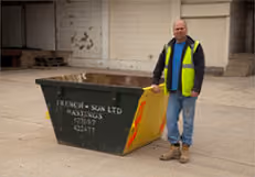 A man in a high-visibility vest stands beside a large green construction skip.