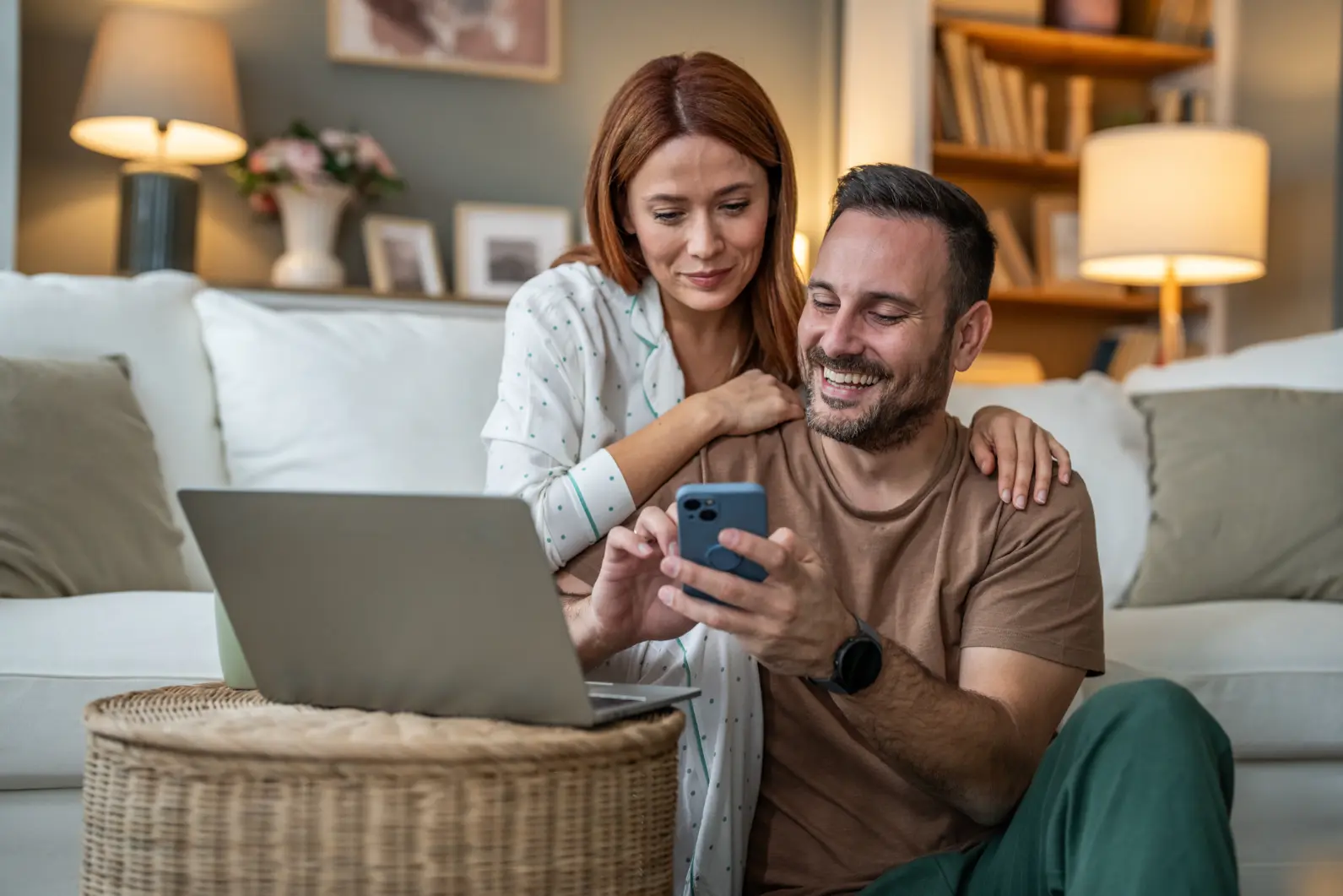 Smiling couple sitting in living room, man holding smartphone, woman leaning on his shoulder, laptop nearby.