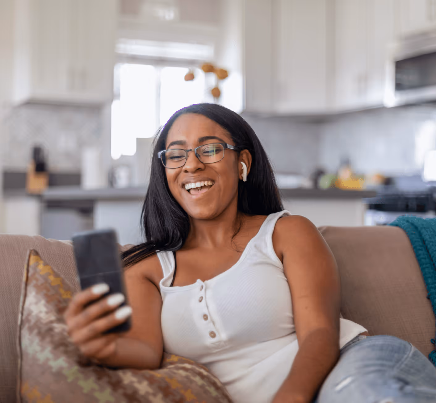Smiling woman with glasses and earbuds relaxing on couch and holding smartphone at home.