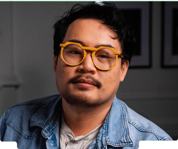Man wearing yellow round glasses looking thoughtfully toward the camera in a room with art on the wall in the background.
