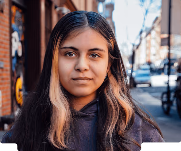 Young woman with long dark hair featuring blonde streaks, wearing a dark hoodie, standing outdoors on a city street.