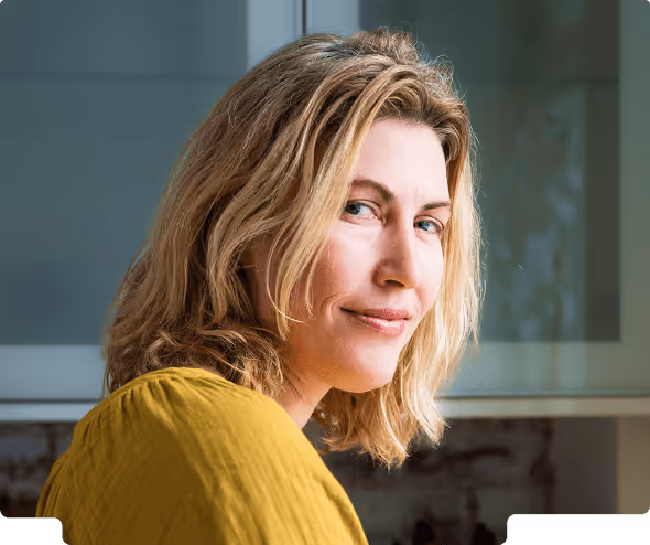 Woman with shoulder-length blonde hair wearing a mustard yellow top, smiling slightly and looking over her shoulder indoors.