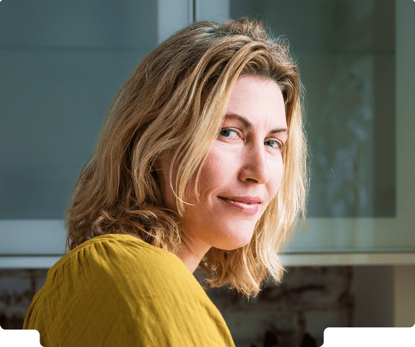 Woman with shoulder-length blonde hair wearing a mustard yellow top, smiling slightly and looking over her shoulder indoors.