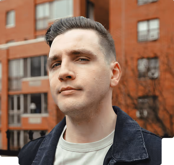 Young man with short dark hair wearing a dark jacket and light shirt standing in front of a red brick building.