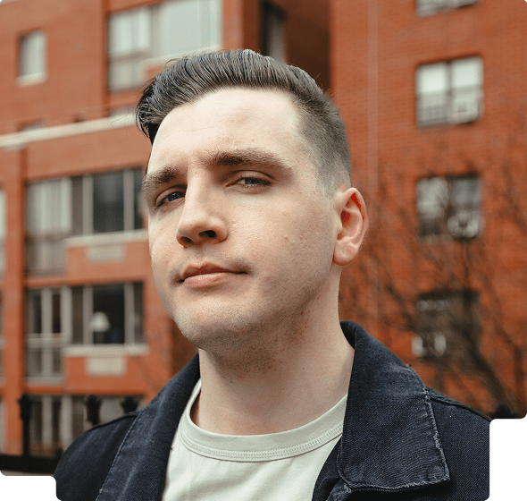 Young man with short dark hair wearing a dark jacket and light shirt standing in front of a red brick building.