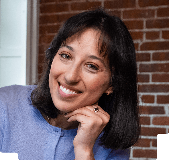 Smiling woman with shoulder-length dark hair wearing a light purple sweater, resting her chin on her hand against a background of a brick wall.