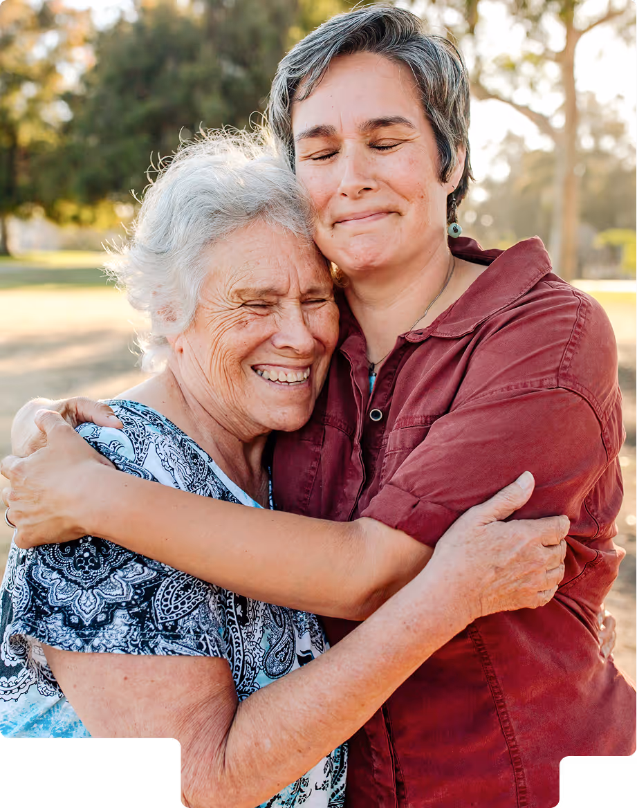 Two women embracing outdoors with eyes closed and smiling, expressing warmth and affection.