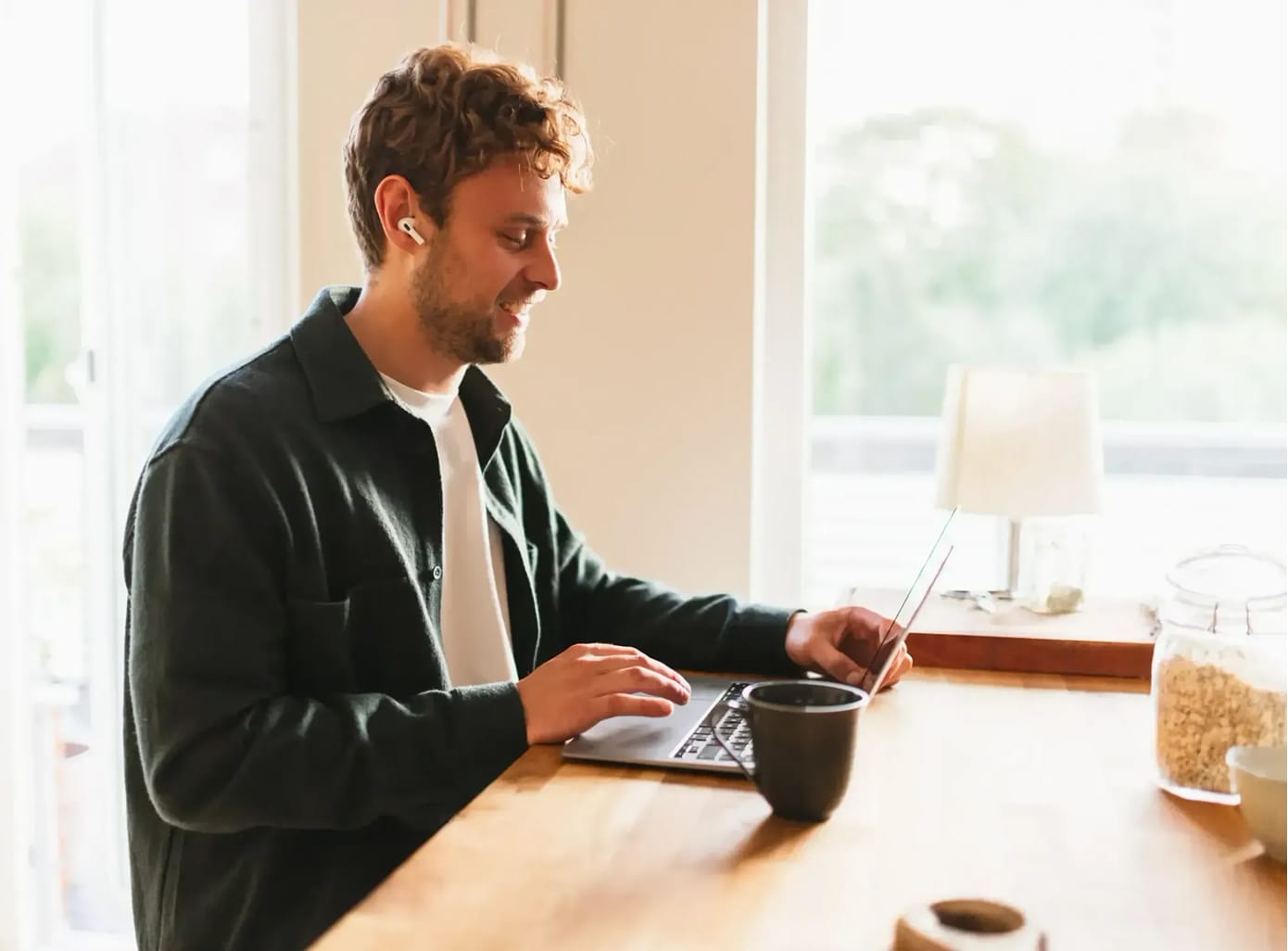Man with earbuds smiling and working on a laptop at a wooden table with a mug nearby.