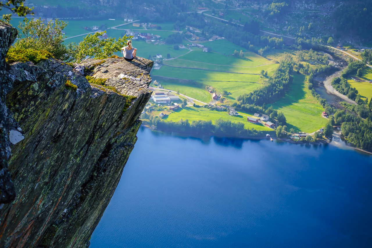 Person sitting on cliff edge overlooking green fields, houses, and a blue fjord below.