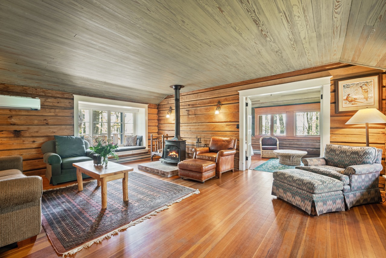 Cozy living room with wood-paneled walls and ceiling, a fireplace, leather and patterned armchairs, a wooden coffee table, and a window seat with cushions.