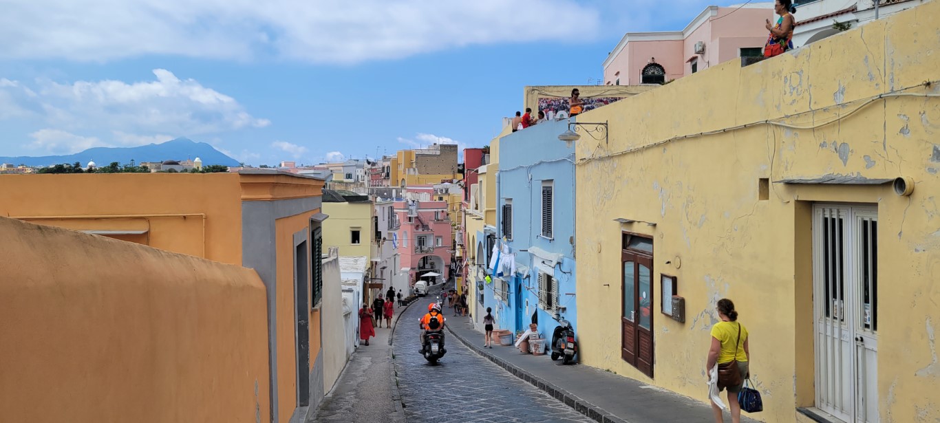 A street view in Procida, Italy of brightly painted homes and people walking and driving scooters.