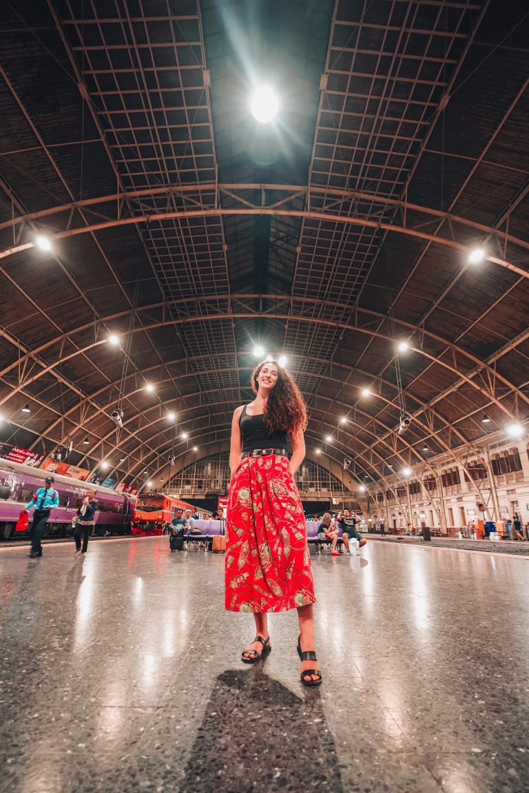A woman wearing a long red skirt posing for the camera at a big, open train station