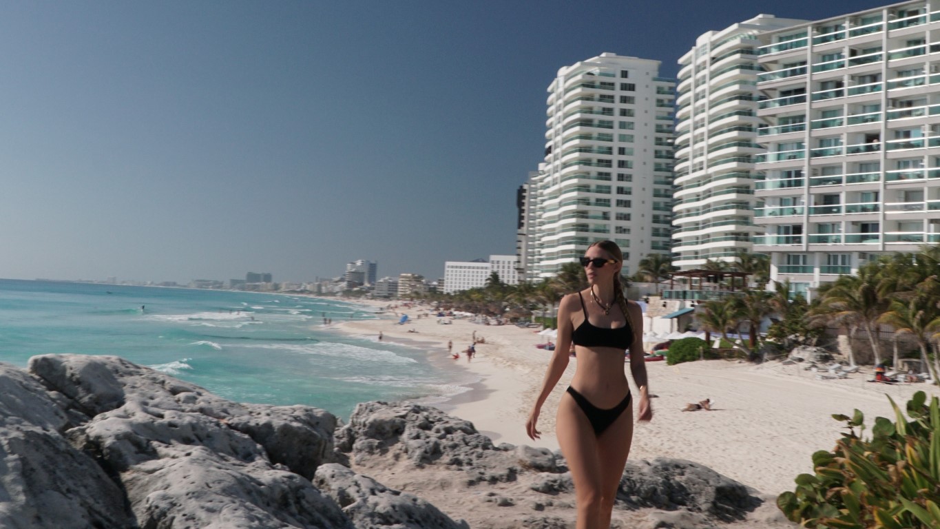 A woman living in Cancun walking towards the camera in a black bikini on the beach in front of a row of hotels