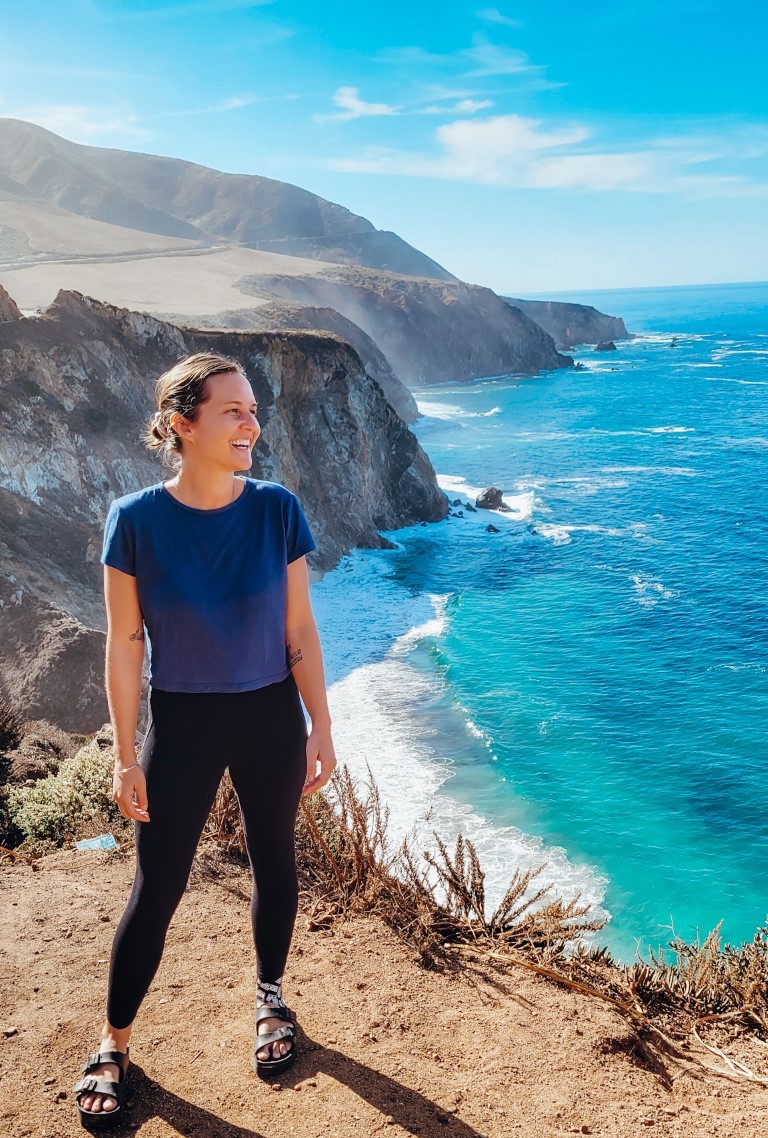 a woman smiling on the edge of a viewpoint off Hwy 1 in California with a rocky coast behind her