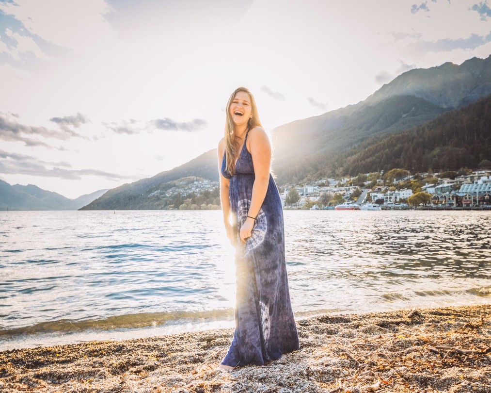 A female expat in New Zealand o the working holiday visa standing at the shore of a lake laughing into the sunlight