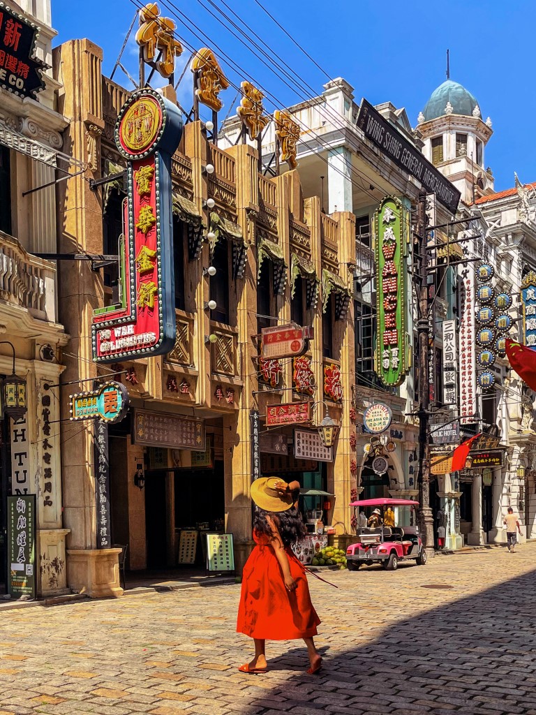 a woman running across the street, wearing a red dress, holding her hat, in a town that looks like a movie set in china