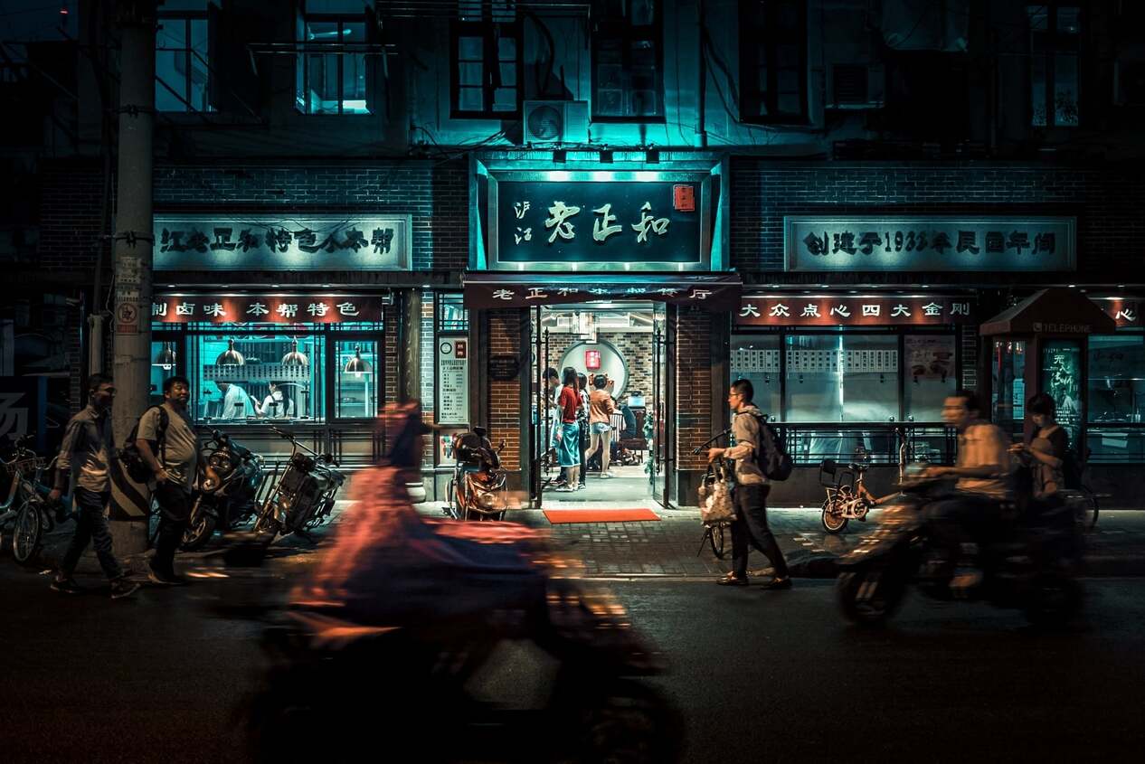 A shot of a Chinese store at night illuminated slightly with motorbikes buzzing past