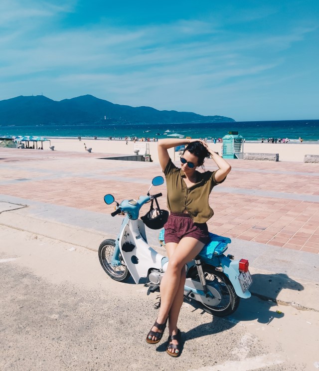 A foreign woman sitting on her motorbike on the coast in Vietnam with the beach and mountains behind her
