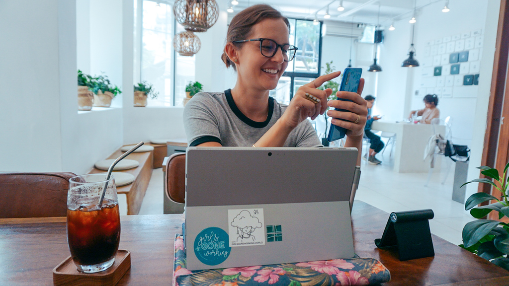 A woman at a coffee shop using her phone with her computer open in front of her and a coffee