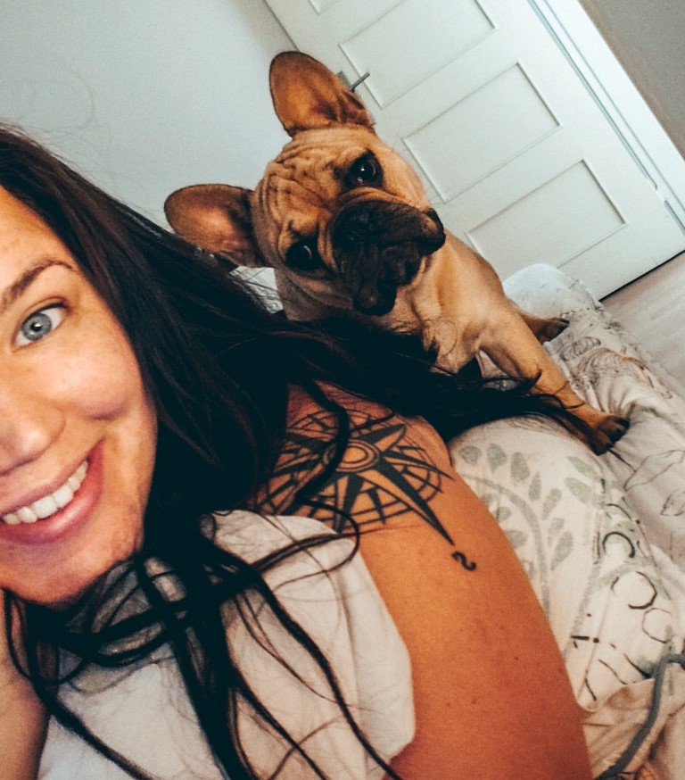 A woman taking a selfie while laying on the bed with a pug puppy looking directly at the cameraa