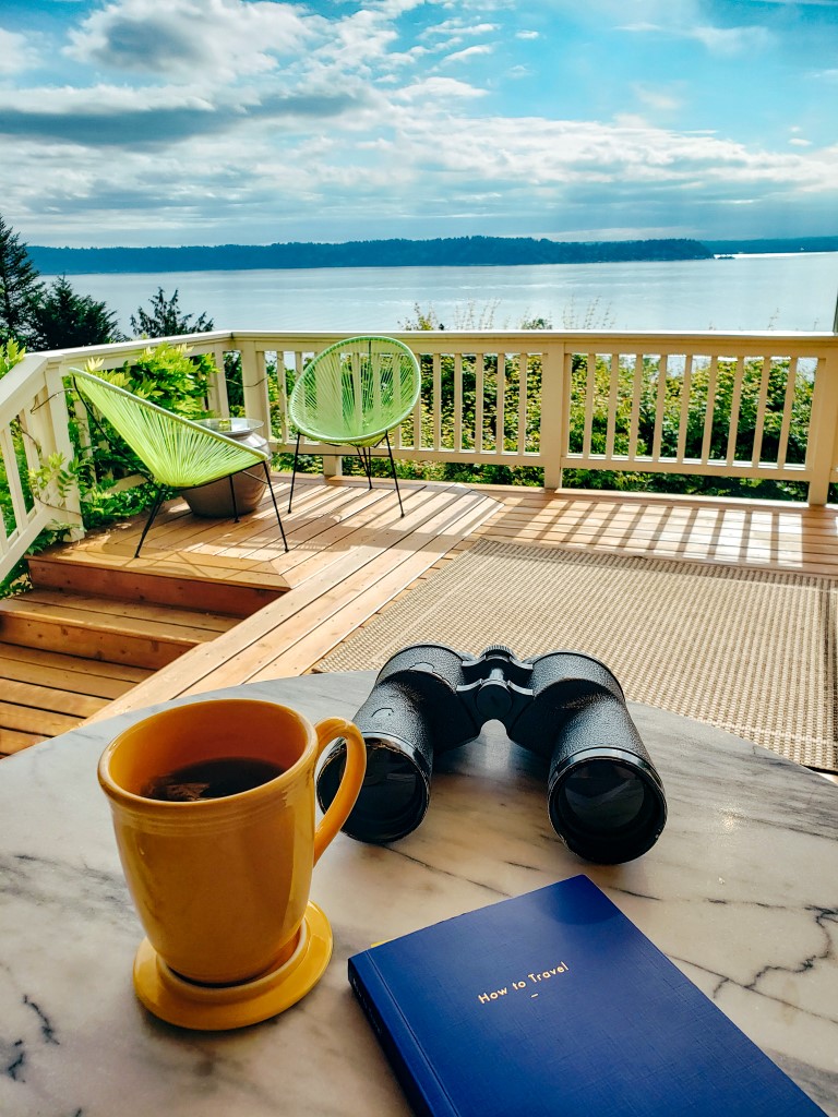 a beautiful view of a lake on a sunny day with a pair of binoculars, a small travel book and a coffee on a table outside