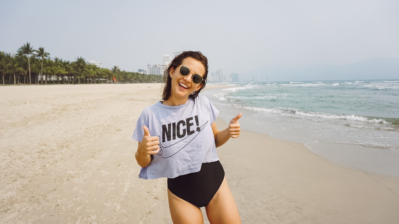 woman on a beach in Vietnam wearing a one piece bathing suit and t-shirt that says "nice" giving two thumbs up to the camera
