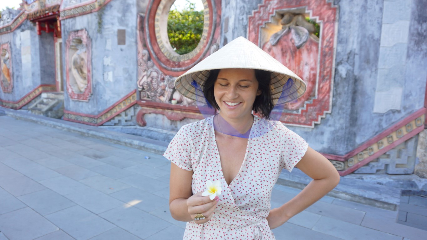 A foreign woman wearing a conical hat looking at a flower at a temple in Hoi An, Vietnam