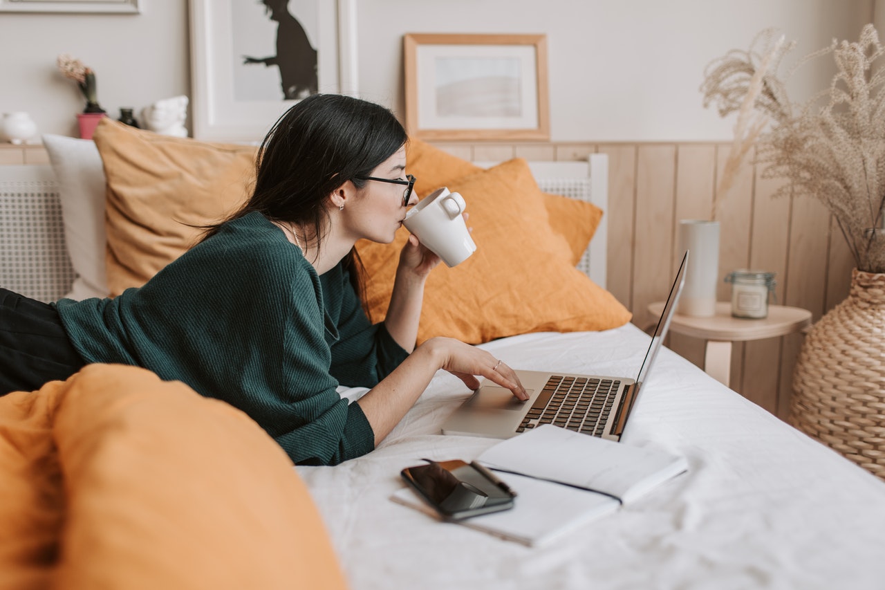 A woman working comfortably from bed while drinking a coffee and browsing her computer