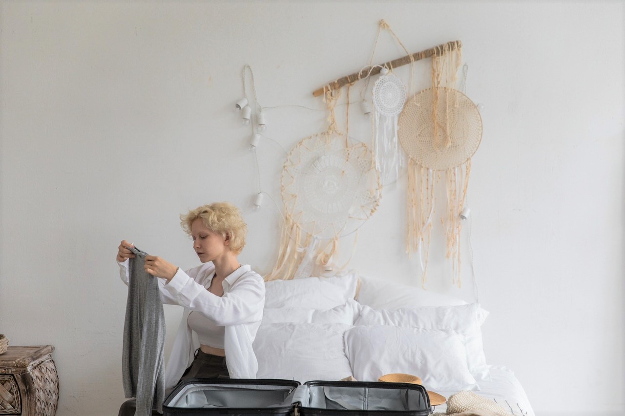 A blonde woman sitting on her bag, folding clothes to pack into a suitcase