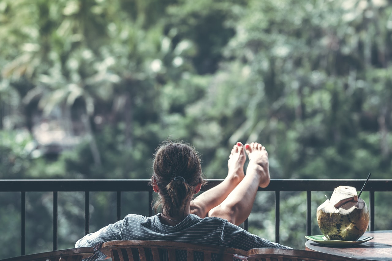 A relaxed woman drinking a coconut looking out into the jungle while lounging on a chair with her feet propped up