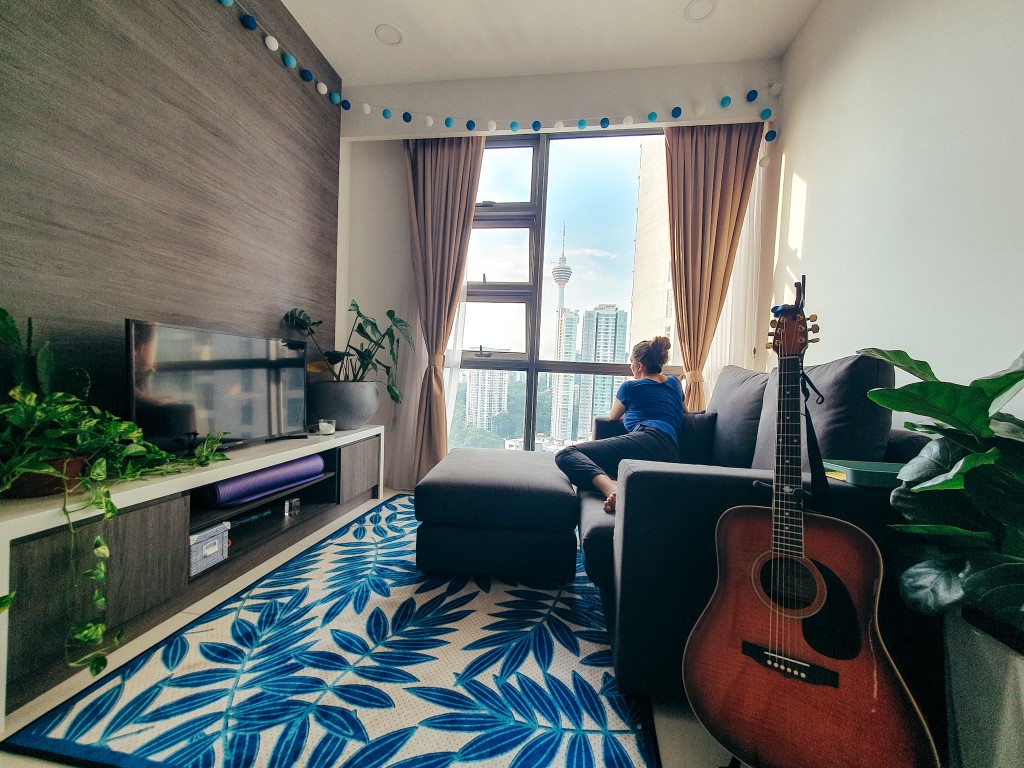 the living room of an apartment in a high rise building in Kuala Lumpur with a TV, coach, coffee table, guitar and a woman looking out the window at the view