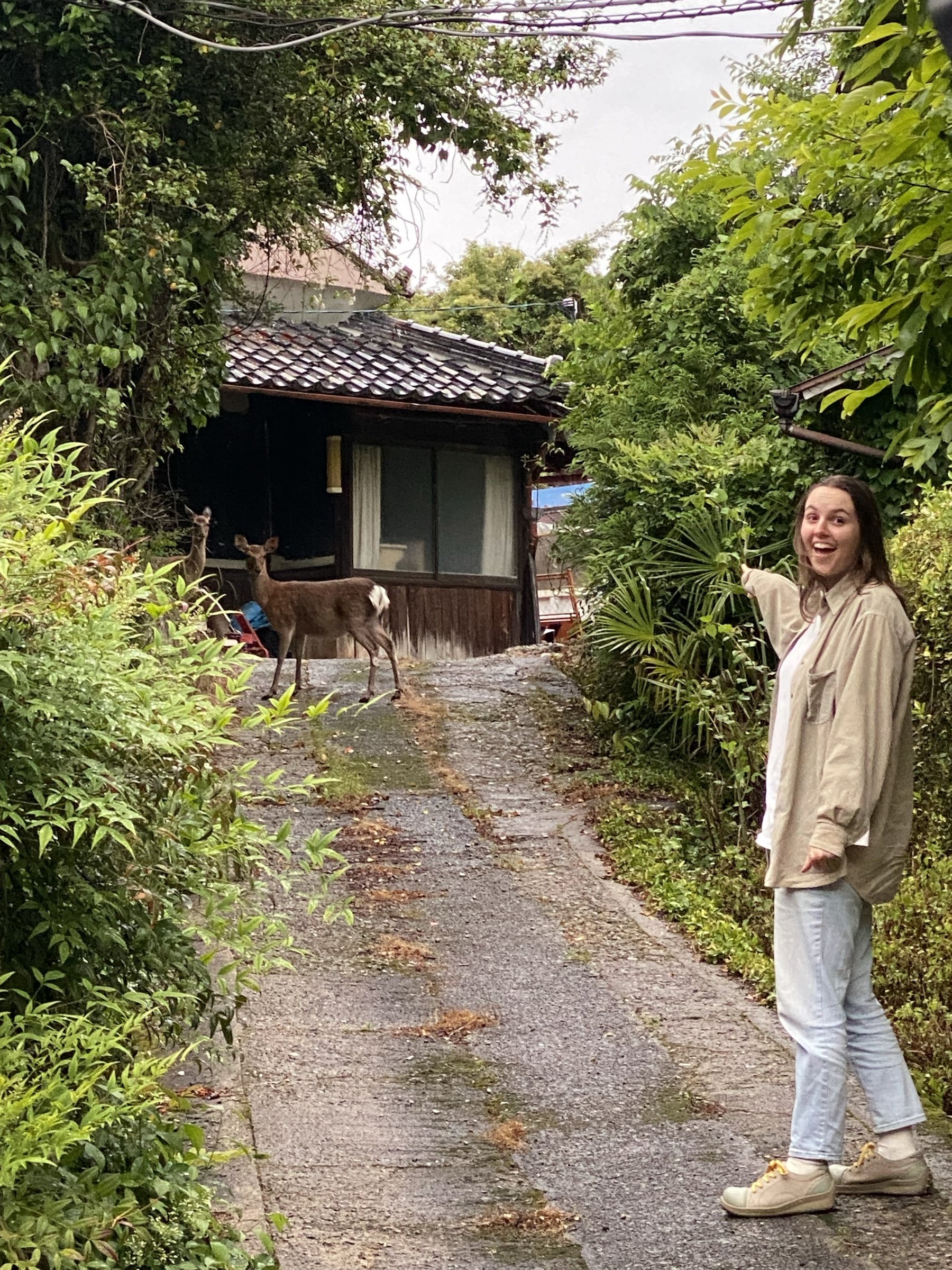 Robyn pointing at two deer standing in a narrow lane beside an old wooden house.